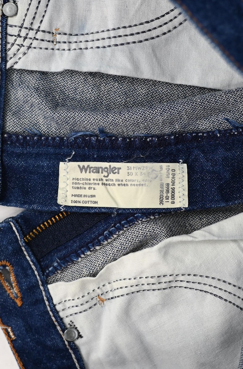 以下が含まれることがあります： Wranglerのジーンズの内側のクローズアップ。ラベルには「Wrangler 31 MWZW 30 x 34」と「Made in USA 100% Cotton」と記載されています。ジーンズは、白いステッチとジッパーが付いた濃い青色のデニムです。