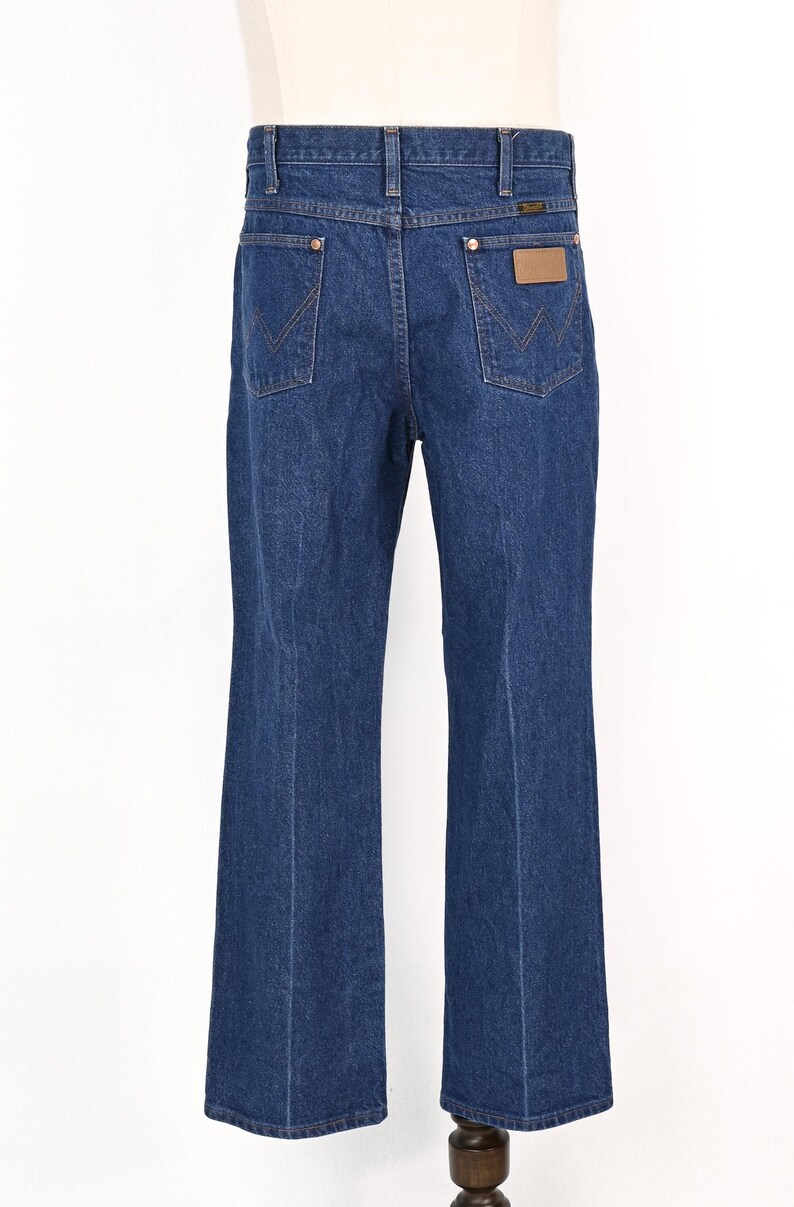 May include: Dark blue denim jeans with a classic five-pocket design. Features a brown leather patch and the Wrangler logo on the back pocket. The jeans have a straight leg and are displayed on a mannequin.