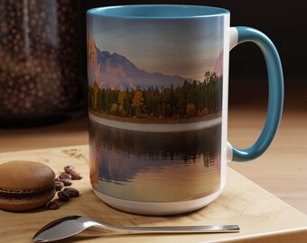 May include: A white ceramic mug with a teal rim and handle. The mug features a scenic image of a mountain range reflected in a lake at sunset.