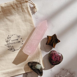 May include: A set of five crystals in a white cotton drawstring bag. The crystals are a pink rose quartz point, a green aventurine heart, a brown tiger's eye star, a pink and green rhodochrosite heart, and a green and black emerald crystal. The bag has the text "Eclectic Earth Finds" printed on it.