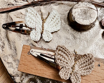 May include: Two crocheted butterfly hair clips. One is white and the other is beige. Both have gold accents.