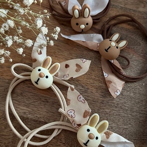 Puede incluir: Tres gomas para el pelo con dijes en forma de conejo. Las gomas para el pelo son marrones, beige y blancas. Los dijes de conejo son marrones, blancos y beige. Las gomas para el pelo están decoradas con lazos de tela marrones y blancos.