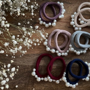 Può includere: Un set di otto scrunchies per capelli con decorazioni di perle. Gli scrunchies sono di vari colori, tra cui bordeaux, blu navy, grigio e beige.