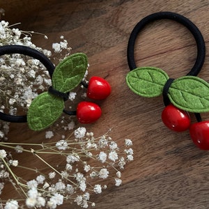 Könnte beinhalten: Zwei schwarze Haargummis mit roten Filz-Kirschen und grünen Filz-Blättern.