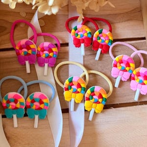 May include: A collection of hair ties featuring colorful lollipop charms with bows. The lollipops are multi-colored with swirls of pink, yellow, blue, and green. Each hair tie has a matching elastic band in pink, red, light blue, or yellow.