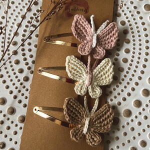 May include: Three crocheted butterfly hair clips on a brown background. The butterflies are in shades of pink, white, and brown. The hair clips are gold.