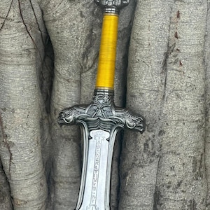 May include: A detailed image of a decorative sword. The sword features a silver-colored blade and crossguard with intricate carvings. The handle is wrapped in bright yellow material. The pommel has a skull design.