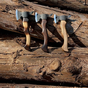 May include: Three axes with wooden handles and metal blades are displayed against a textured wooden background. The handles have a curved design, and the blades have a silver finish. The wood grain on the handles varies in color.