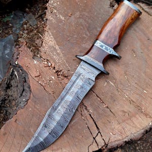 May include: A hunting knife with a brown wooden handle and a patterned steel blade. The knife is resting on a wooden surface.