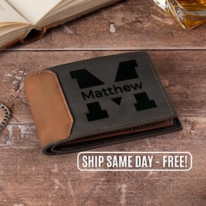 May include: A personalized bi-fold wallet with the name "Matthew" and a large letter "M" engraved on the front. The wallet features a gray body with a tan accent and is made of leather.