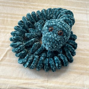May include: A teal crocheted snake toy. The snake is coiled, with a rounded head and two brown eyes. The body is made of soft, textured yarn, creating a cuddly appearance. The toy is designed to be a child's plaything.