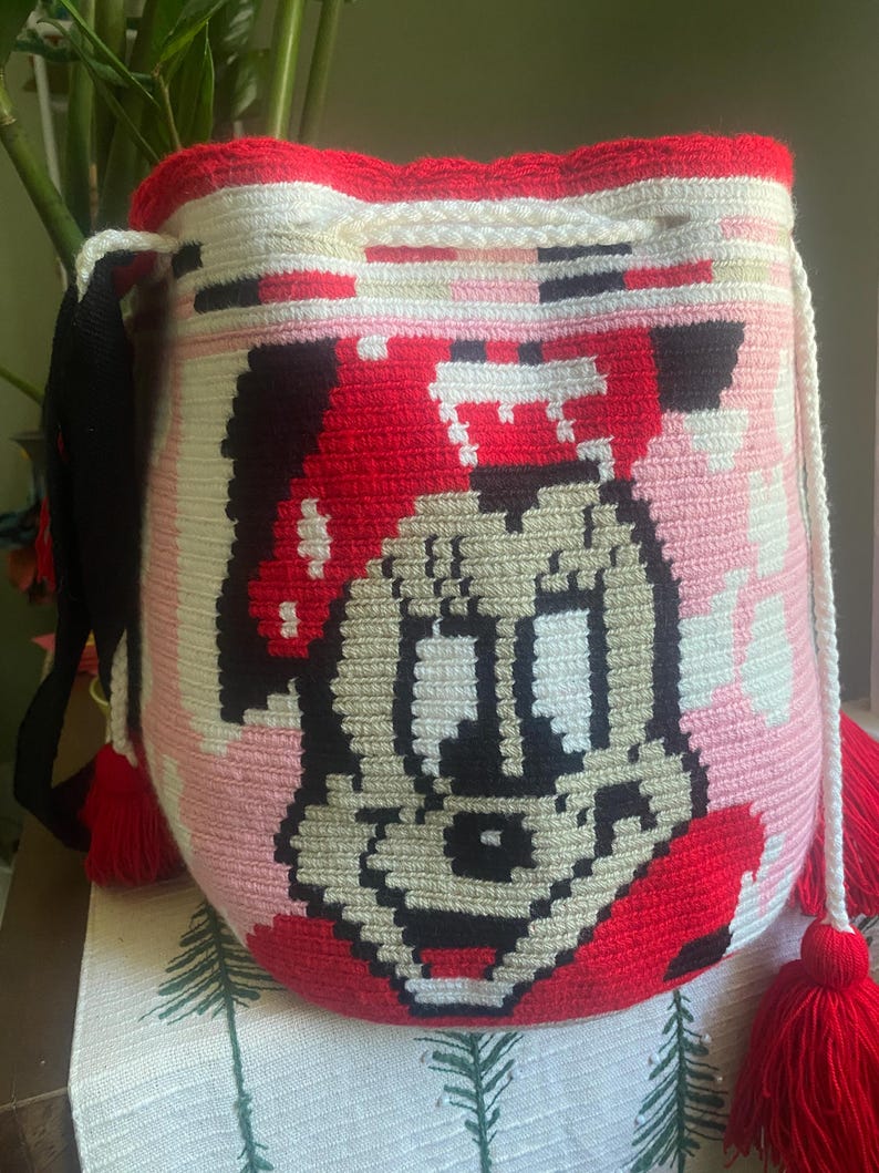 Puede incluir: Un bolso tipo saco de ganchillo cil&iacute;ndrico con un gr&aacute;fico pixelado de un rat&oacute;n de dibujos animados con un gorro rojo. El bolso est&aacute; hecho con hilo blanco, rojo, negro y beige, incluye un cierre de cord&oacute;n blanco y borlas de hilo rojo.