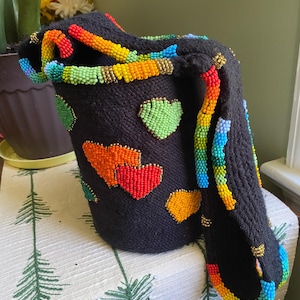 May include: A black bucket-style bag featuring a knitted texture and decorated with colorful beaded heart motifs in shades of red, orange, and green. The bag includes a wide shoulder strap adorned with a vibrant rainbow-colored beaded trim.