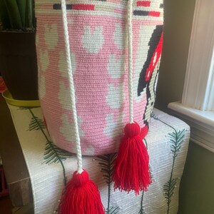 Puede incluir: Un bolso tipo cubo de ganchillo cil&iacute;ndrico con una base rosa claro y un patr&oacute;n blanco repetitivo. El borde superior est&aacute; acentuado con una banda roja vibrante y un asa negra. Dos cordones trenzados blancos cuelgan, cada uno terminado en una gran borla roja.