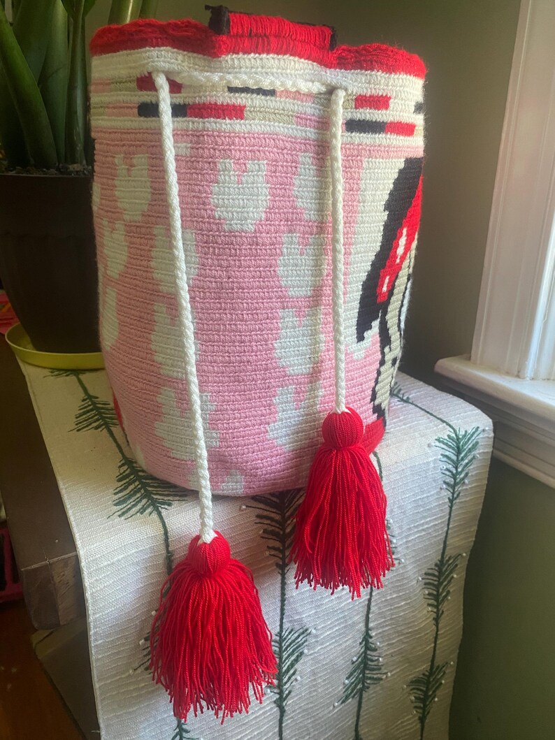 Puede incluir: Un bolso tipo cubo de ganchillo cil&iacute;ndrico con una base rosa claro y un patr&oacute;n blanco repetitivo. El borde superior est&aacute; acentuado con una banda roja vibrante y un asa negra. Dos cordones trenzados blancos cuelgan, cada uno terminado en una gran borla roja.