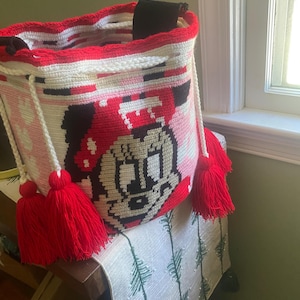 Puede incluir: Un bolso tipo cubo de ganchillo cil&iacute;ndrico con un gr&aacute;fico de un rat&oacute;n de dibujos animados con un lazo rojo. El bolso es principalmente blanco con detalles en rojo y negro, un borde de ganchillo rojo y dos cordones blancos con borlas de hilo rojo.