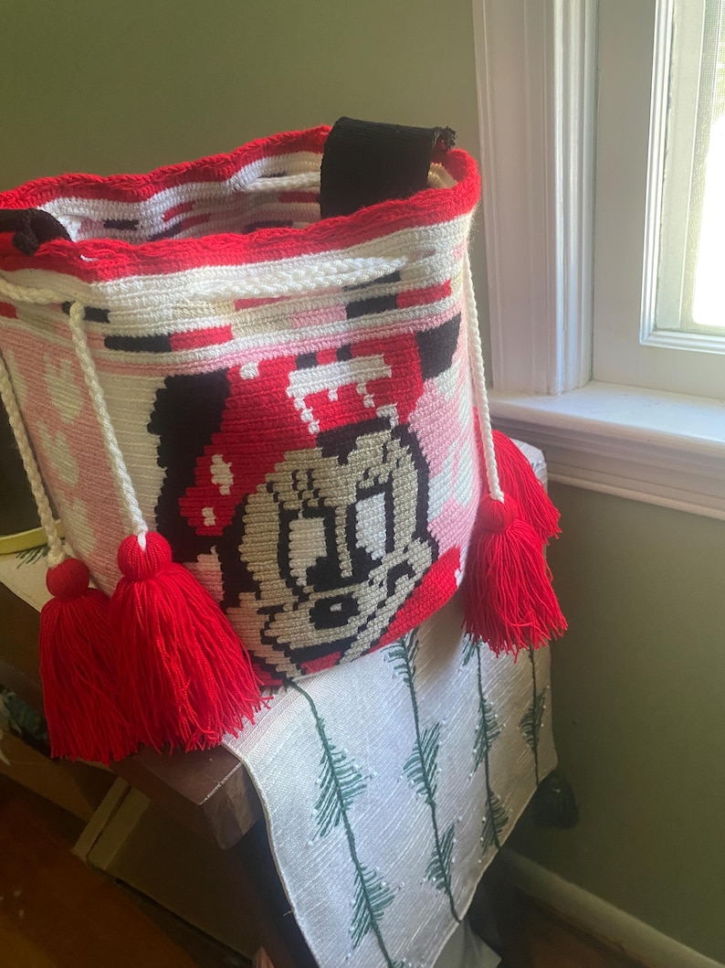 Puede incluir: Un bolso tipo cubo de ganchillo cil&iacute;ndrico con un gr&aacute;fico de un rat&oacute;n de dibujos animados con un lazo rojo. El bolso es principalmente blanco con detalles en rojo y negro, un borde de ganchillo rojo y dos cordones blancos con borlas de hilo rojo.