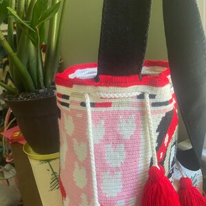Puede incluir: Un bolso tipo cubo de ganchillo cil&iacute;ndrico con un patr&oacute;n de corazones rosas y blancos, un borde superior rojo s&oacute;lido y una correa de hombro negra ancha. El bolso incluye un cierre de cord&oacute;n trenzado blanco con tres borlas de hilo rojo.