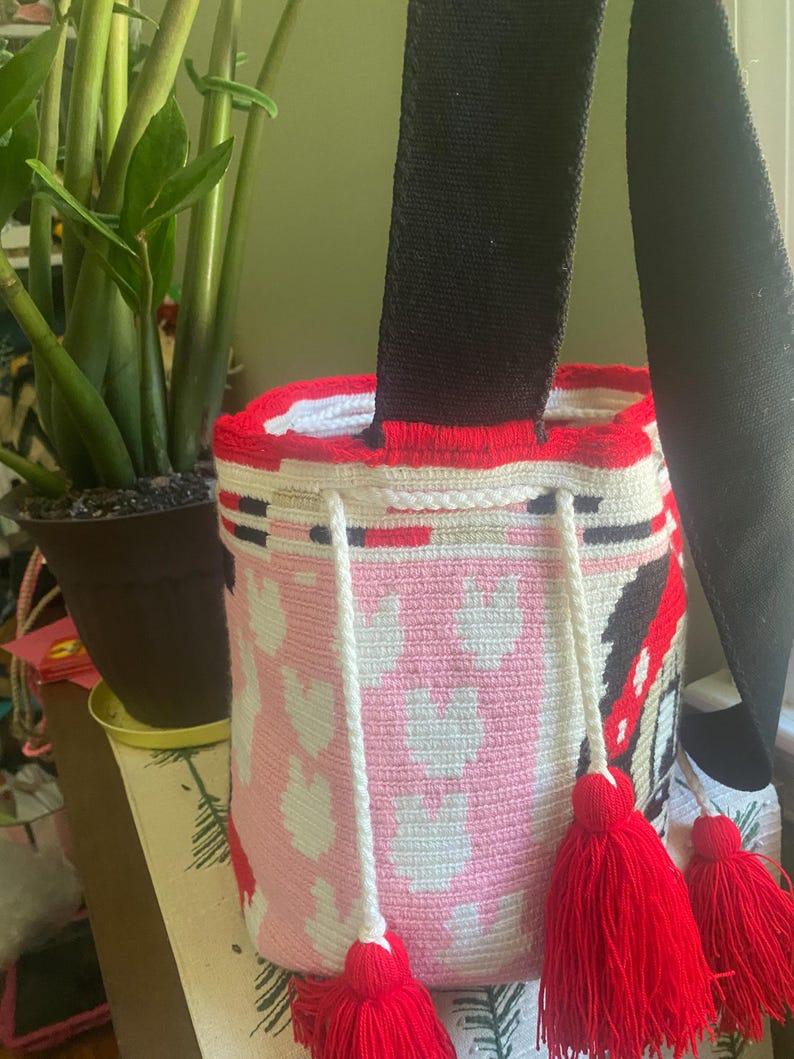 Puede incluir: Un bolso tipo cubo de ganchillo cil&iacute;ndrico con un patr&oacute;n de corazones rosas y blancos, un borde superior rojo s&oacute;lido y una correa de hombro negra ancha. El bolso incluye un cierre de cord&oacute;n trenzado blanco con tres borlas de hilo rojo.