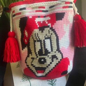 Puede incluir: Un bolso tipo cubo de ganchillo cil&iacute;ndrico con un gr&aacute;fico pixelado de una cara de rat&oacute;n de dibujos animados en negro, blanco y beige. El bolso tiene un borde superior rojo vibrante, un cierre de cord&oacute;n blanco con dos borlas rojas grandes y una correa negra ancha.