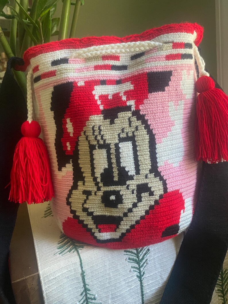 Puede incluir: Un bolso tipo cubo de ganchillo cil&iacute;ndrico con un gr&aacute;fico pixelado de una cara de rat&oacute;n de dibujos animados en negro, blanco y beige. El bolso tiene un borde superior rojo vibrante, un cierre de cord&oacute;n blanco con dos borlas rojas grandes y una correa negra ancha.