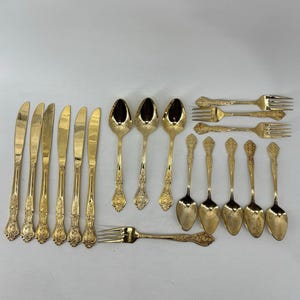 Puede incluir: Un conjunto de cubiertos dorados, que incluye cuchillos, cucharas y tenedores, dispuestos sobre un fondo claro. Los utensilios tienen detalles florales ornamentados en los mangos. El juego incluye varios tamaños de cucharas, tenedores y cuchillos.