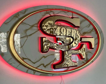 Puede incluir: Un logotipo de los San Francisco 49ers en oro y rojo con un fondo de mármol. El logotipo está rodeado por un halo rojo. El logotipo presenta el nombre del equipo y el número 49ers.