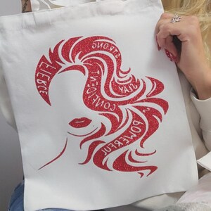 May include: White tote bag featuring a red glitter design of a woman's silhouette and the words "Some Kind of Wonderful" and "Fierce". The bag has white handles and is made of a durable fabric.