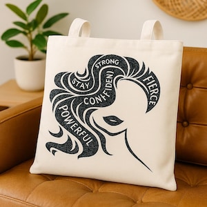 May include: Cream-colored tote bag with black graphic of a woman's silhouette and flowing hair. The words "Stay, Confident, Powerful, Strong, Fierce" are incorporated into the design. The bag has two cream-colored handles.