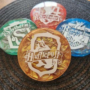 Puede incluir: Cuatro posavasos redondos de resina, coloridos, con los escudos de las casas de Hogwarts: Gryffindor (rojo), Ravenclaw (azul), Hufflepuff (dorado) y Slytherin (verde). Cada posavasos tiene un emblema y texto blanco.
