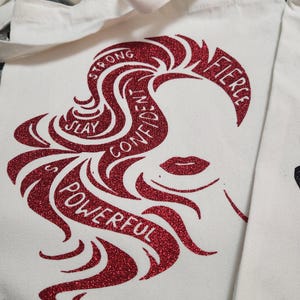 May include: White canvas tote bag with a red glitter design of a woman's silhouette and the words "Strong, Slay, Confident, Powerful, Fierce". The tote bag has white handles.