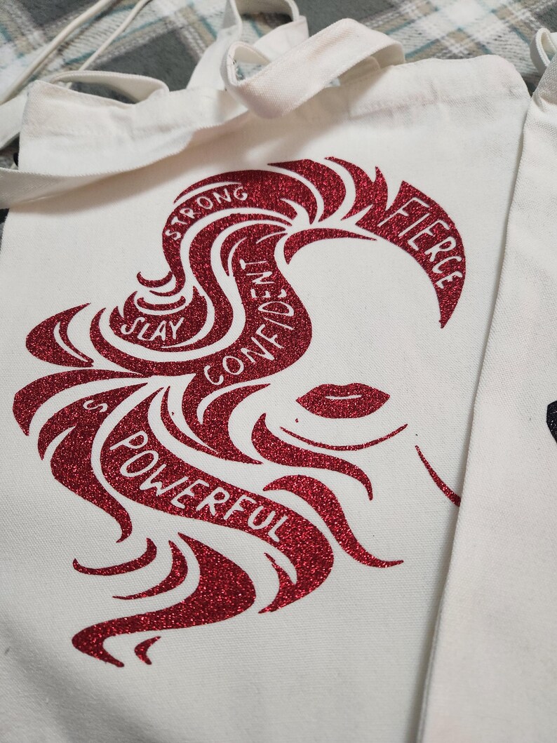 May include: White canvas tote bag with a red glitter design of a woman's silhouette and the words "Strong, Slay, Confident, Powerful, Fierce". The tote bag has white handles.