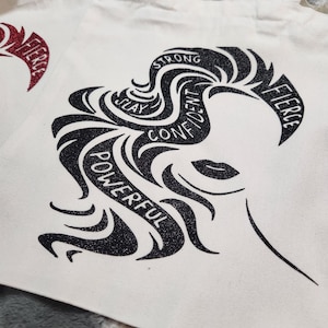 May include: White canvas tote bag featuring a black and glittery graphic of a woman's silhouette with the words "Strong, Slay, Confident, Powerful, Fierce" incorporated into the design. The tote bag has two handles.