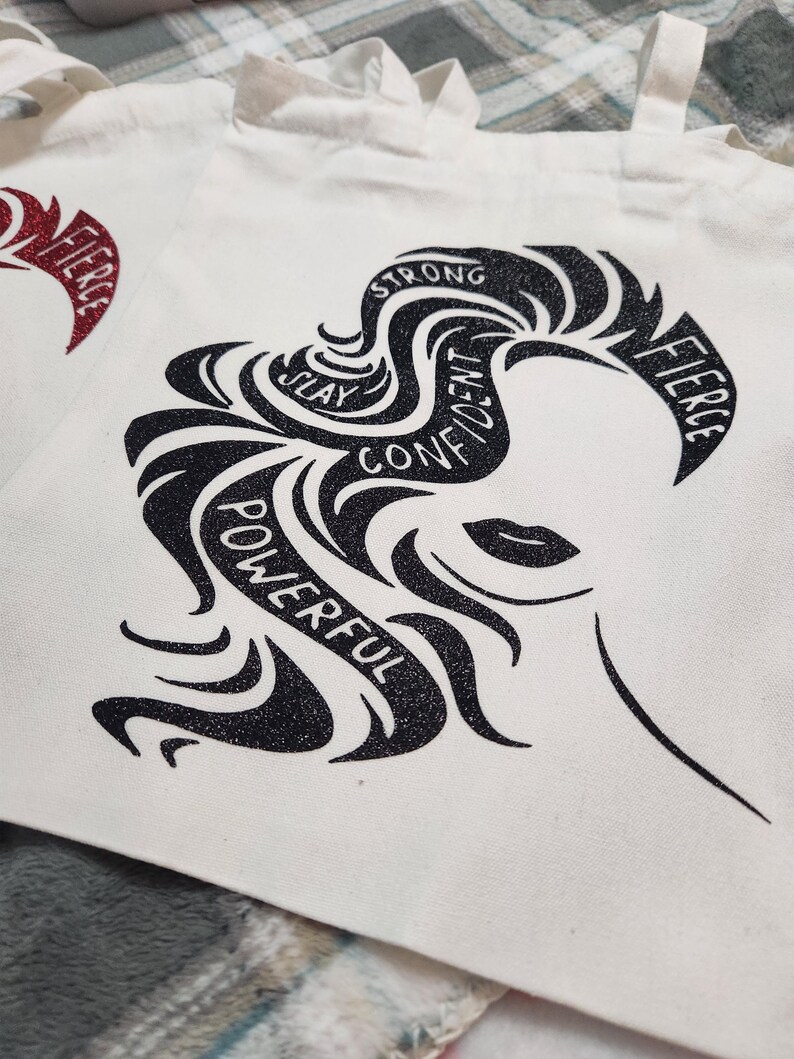May include: White canvas tote bag featuring a black and glittery graphic of a woman's silhouette with the words "Strong, Slay, Confident, Powerful, Fierce" incorporated into the design. The tote bag has two handles.