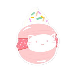 May include: A pink macaron with a white cat face peeking out from the center. The macaron is topped with a swirl of white frosting and colorful sprinkles.