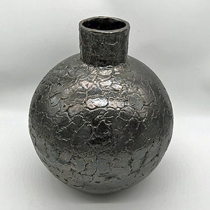 Può includere: Un vaso in ceramica grigio scuro con una superficie strutturata e screpolata. Il vaso ha un corpo rotondo e un collo cilindrico. L'aspetto artigianale del vaso lo rende adatto per esporre fiori o come elemento decorativo. Un elegante oggetto d'arredo.