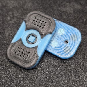 May include: Two-piece, 3D-printed fidget toy in light blue and black. The top piece has a central star-shaped design and perforated sections. The bottom piece is light blue with a circular pattern and a screw.