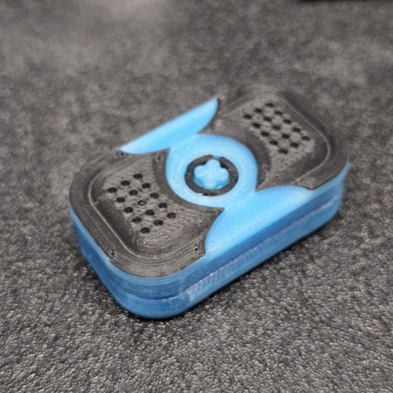 May include: A 3D-printed, rectangular object with a blue base and black top. The top features a series of small holes and a central circular design with a four-leaf clover shape. The object is likely a case or container.
