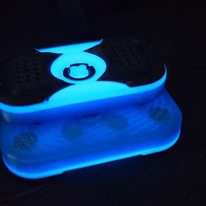 May include: A glowing blue and black electronic device with a rectangular shape. The device has a rounded top with a central design and speaker-like features. The bottom section also glows blue, creating a striking visual effect.