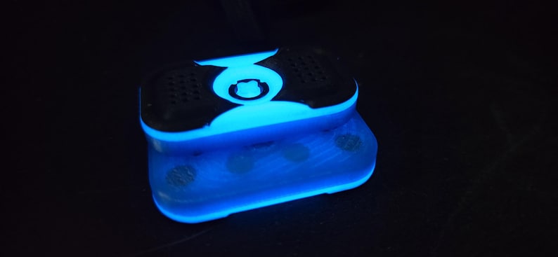 May include: A glowing blue and black electronic device with a rectangular shape. The device has a rounded top with a central design and speaker-like features. The bottom section also glows blue, creating a striking visual effect.