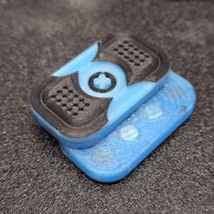 May include: A two-piece, 3D-printed object in blue and black. The top piece has a black, perforated design and a central cross-shaped detail. The bottom piece is a solid blue rectangle. The object is likely a component or accessory.