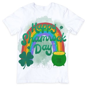 Op de afbeelding: Wit T-shirt met een groen klavertjevier en een regenboogontwerp. De tekst "Happy Shamrock Day" staat in het groen op het shirt.