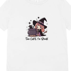 May include: White t-shirt with a cartoon illustration of a witch stirring a cauldron with a black cat beside her. The text "Too Cute To Spook" is printed below the image.