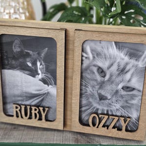 May include: A wooden double photo frame with two black and white cat portraits. The frame has the names "RUBY" and "OZZY" cut out below each photo. The frame is made of light-colored wood.