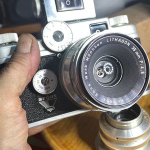 Könnte beinhalten: Eine silberne und schwarze 35-mm-Filmkamera mit einem Lithagon 35-mm-Objektiv f:4.5. Die Kamera hat eine schwarze Lederabdeckung und ein silbernes Metallgehäuse. Das Objektiv hat einen schwarzen Ring mit weißer Beschriftung, auf dem "Enna Werk München Lithagon 35mm F:4.5" steht. Die Kamera hat ein silbernes Metallzifferblatt mit einem schwarzen Ring, auf dem "FEET" steht.