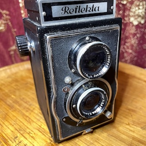May include: A black and silver vintage camera with the name "Reflekta" on the top. The camera has two lenses, one with a black and silver ring and the other with a black ring. The camera is sitting on a wooden surface.