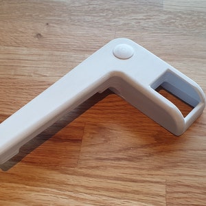 Könnte beinhalten: Weiße Eckverbindung aus Kunststoff mit einem runden Knopf oben. Die Verbindung ist so konzipiert, dass sie die Ecken von Möbeln oder anderen Gegenständen sichert.