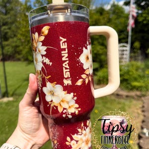 Puede incluir: Un vaso Stanley rojo brillante con un asa y tapa color crema. El vaso est&aacute; decorado con dise&ntilde;os florales blancos y amarillos. El logotipo de Stanley es visible en el lateral. El vaso est&aacute; sostenido por una persona.