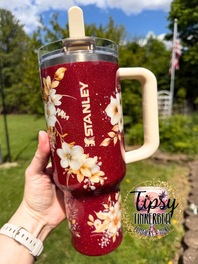 Puede incluir: Un vaso Stanley rojo brillante con un asa y tapa color crema. El vaso est&aacute; decorado con dise&ntilde;os florales blancos y amarillos. El logotipo de Stanley es visible en el lateral. El vaso est&aacute; sostenido por una persona.