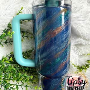 Puede incluir: Un vaso grande y aislado con un asa y tapa de color turquesa. El vaso presenta un dise&ntilde;o arremolinado de purpurina azul, turquesa y dorada. Las palabras "Tipsy Tinkerbell Tumblers & More" est&aacute;n impresas en la parte inferior.